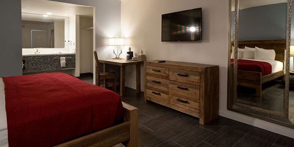 A hotel room with a king-size bed, wooden dresser, desk, flat-screen TV, and bathroom in the background.