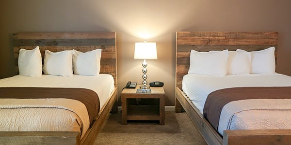 A hotel room with two double beds, wooden headboards, a nightstand with a lamp, and a carpeted floor.