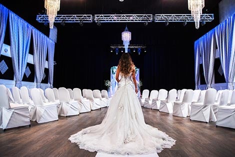 A woman in a wedding dress standing in a large event space with white chairs and blue lighting.
