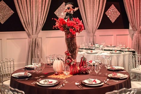 A round dining table set with a brown tablecloth, plates, glasses, and a floral centerpiece with pumpkins.