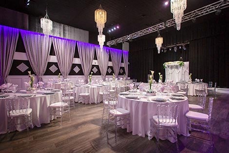 A large, elegantly set dining room with round tables, white tablecloths, clear chairs, and chandeliers.