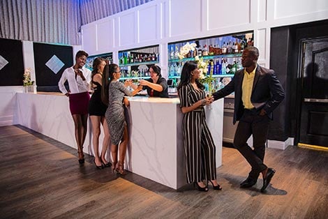 A group of people socializing at a modern bar with a marble countertop and shelves with bottles.