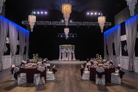 A formal dining room with tables set for a large event, featuring chandeliers, a stage, and a backdrop.